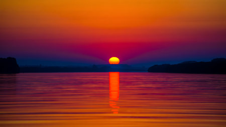 A breathtaking sunset descends over a tranquil river, casting a golden-orange glow across the horizon and reflecting vividly on the calm water surface. Dark silhouettes of trees frame the scene, creating a serene contrast to the warm gradient sky that shifts from deep orange to violet. This peaceful moment captures the natural beauty and stillness of twilight.の素材