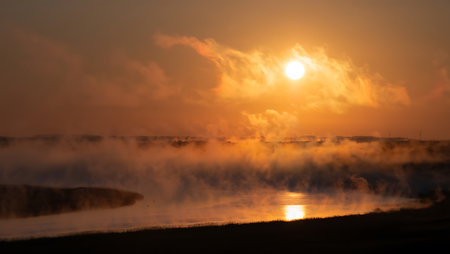 A stunning sunset radiates golden and fiery hues over a winding river, with mist rising gently above the water. The brilliant sun hovers above the tree-lined horizon, casting a warm orange glow across the landscape. Wisps of fog and rich reflections add depth and drama, evoking a mystical and tranquil atmosphere in the natural setting.の素材