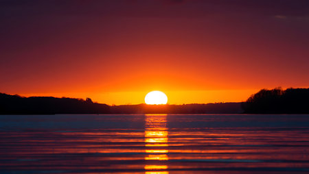 A breathtaking sunset descends over a tranquil river, casting a golden-orange glow across the horizon and reflecting vividly on the calm water surface. Dark silhouettes of trees frame the scene, creating a serene contrast to the warm gradient sky that shifts from deep orange to violet. This peaceful moment captures the natural beauty and stillness of twilight.の素材