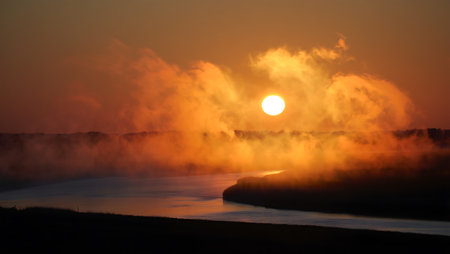 A stunning sunset radiates golden and fiery hues over a winding river, with mist rising gently above the water. The brilliant sun hovers above the tree-lined horizon, casting a warm orange glow across the landscape. Wisps of fog and rich reflections add depth and drama, evoking a mystical and tranquil atmosphere in the natural setting.の素材