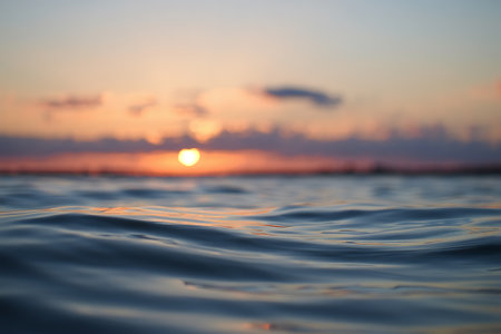 A dreamy sunset casts a golden glow over calm ocean waters, with the sun appearing as a soft blur on the horizon. The low-angle focus captures the gentle ripples and glistening reflections on the water surface, blending warm hues of orange, pink, and purple. This tranquil seascape evokes peace, serenity, and the quiet beauty of nature at dusk.の素材