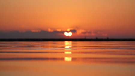 A breathtaking sunset descends over a tranquil river, casting a golden-orange glow across the horizon and reflecting vividly on the calm water surface. Dark silhouettes of trees frame the scene, creating a serene contrast to the warm gradient sky that shifts from deep orange to violet. This peaceful moment captures the natural beauty and stillness of twilight.の素材