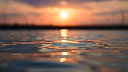 A dreamy sunset casts a golden glow over calm ocean waters, with the sun appearing as a soft blur on the horizon. The low-angle focus captures the gentle ripples and glistening reflections on the water surface, blending warm hues of orange, pink, and purple. This tranquil seascape evokes peace, serenity, and the quiet beauty of nature at dusk.の素材