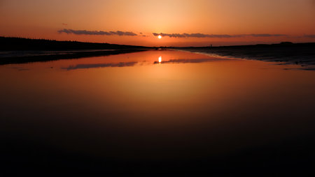 A breathtaking sunset descends over a tranquil river, casting a golden-orange glow across the horizon and reflecting vividly on the calm water surface. Dark silhouettes of trees frame the scene, creating a serene contrast to the warm gradient sky that shifts from deep orange to violet. This peaceful moment captures the natural beauty and stillness of twilight.の素材