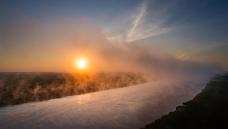 A stunning sunset radiates golden and fiery hues over a winding river, with mist rising gently above the water. The brilliant sun hovers above the tree-lined horizon, casting a warm orange glow across the landscape. Wisps of fog and rich reflections add depth and drama, evoking a mystical and tranquil atmosphere in the natural setting.の素材