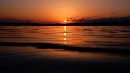 A breathtaking sunset descends over a tranquil river, casting a golden-orange glow across the horizon and reflecting vividly on the calm water surface. Dark silhouettes of trees frame the scene, creating a serene contrast to the warm gradient sky that shifts from deep orange to violet. This peaceful moment captures the natural beauty and stillness of twilight.の素材