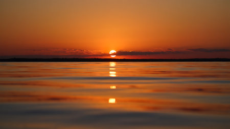 A breathtaking sunset descends over a tranquil river, casting a golden-orange glow across the horizon and reflecting vividly on the calm water surface. Dark silhouettes of trees frame the scene, creating a serene contrast to the warm gradient sky that shifts from deep orange to violet. This peaceful moment captures the natural beauty and stillness of twilight.の素材