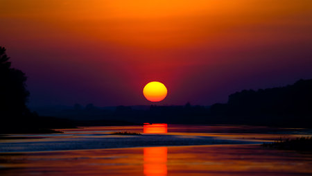 A breathtaking sunset descends over a tranquil river, casting a golden-orange glow across the horizon and reflecting vividly on the calm water surface. Dark silhouettes of trees frame the scene, creating a serene contrast to the warm gradient sky that shifts from deep orange to violet. This peaceful moment captures the natural beauty and stillness of twilight.の素材