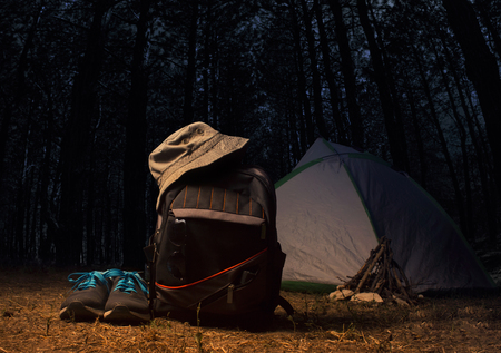 Backpack with travel gear, bucket hat, flashlight, sneakers and travel tent on dark forest night landscape background.の写真素材