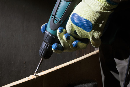 Closeup photo of a worker in outfit with blue gloves using drill with wooden board on black background with upper light.の写真素材