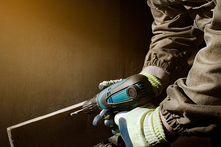 Side view photo of a worker in outfit with blue gloves using drill with wooden board on black background with upper light side view.の写真素材