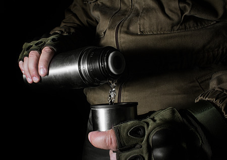 Photo of a male person in brown tactical outfit jacket and gloves pouring out vacuum thermo bottle.の写真素材