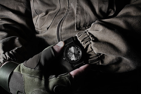 Profile view photo of a male person in brown tactical outfit jacket and gloves holding military watch on black background.の写真素材