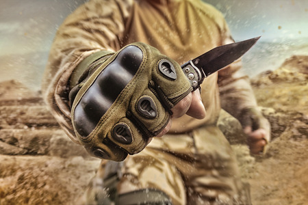 Photo of a military equipped soldier posing and holding a small tactical knife in gloves on desert...