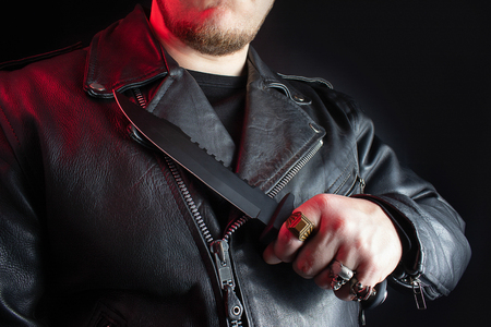 Photo of a biker in black leather jacket with skull rings holding hunting knife on black background.の写真素材