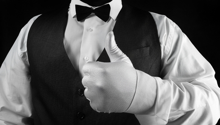 Photo of a waiter in white shirt and black waistcoat showing thumbs up in white glove on black background torso view.の写真素材