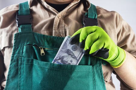 Photo of a worker in green overall outfit with protective gloves pulling out pack of dollar banknotes from a pocket, torso view.の写真素材