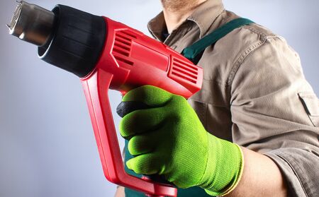 Photo of a worker in green overall outfit with protective gloves holding heat gun tool on grey background torso view.の写真素材