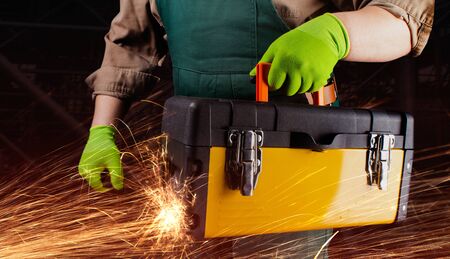 Photo of a worker in green overall outfit with protective gloves holding yellow toolbox with sparks torso view on black construction area background.の写真素材