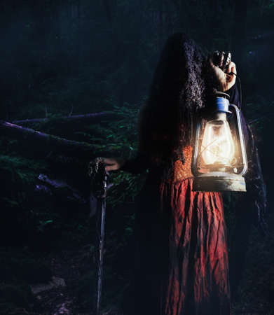Photo of a female witch holding lantern and cane in dark woods gloomy, scary background.の写真素材