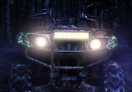Photo of a camouflaged dirty offroad hunting atv vehicle standing at night in forest with headlights turned on, front view.の写真素材