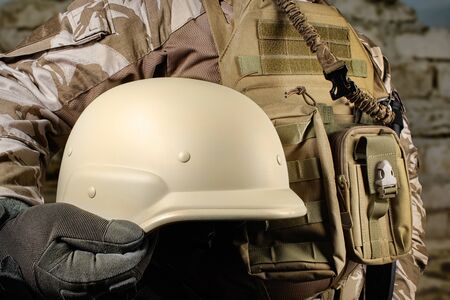 Photo of a fully equipped soldier in uniform, armor holding helmet in desert battlefield background close-up view.の写真素材
