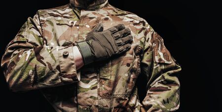 Photo of a soldier in multicam camouflage tunic with hand on a heart on black background.の写真素材