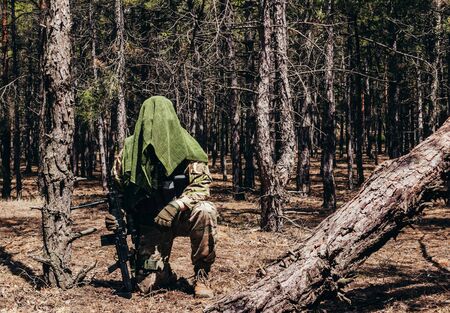 Photo of a fully equipped camouflaged forest sniper with rifle and tactical net scarf sitting in woods.の写真素材