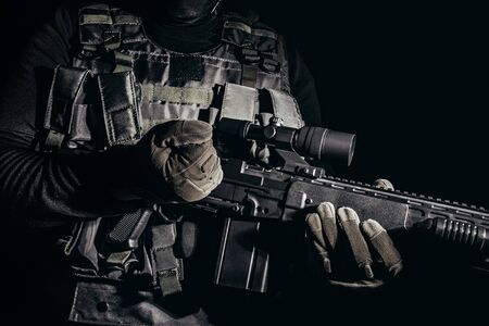 Photo of a soldier in tactical gloves and armor vest standing and reloading sniper rifle on black...