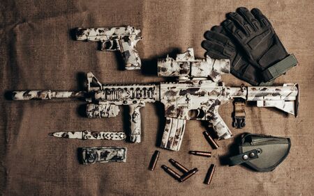 Photo of a camouflaged military AR-15 rifle with scope laying with knife, gun and tactical gloves upper view.の写真素材