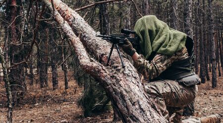 Photo of a fully equipped camouflaged forest sniper with rifle sitting and aiming near the tree in woods.の写真素材