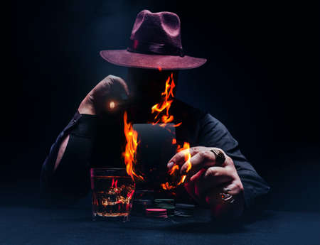 Photo of a demonic gangster with cigar in black shirt and hat playing poker and holding a burning black card.の写真素材
