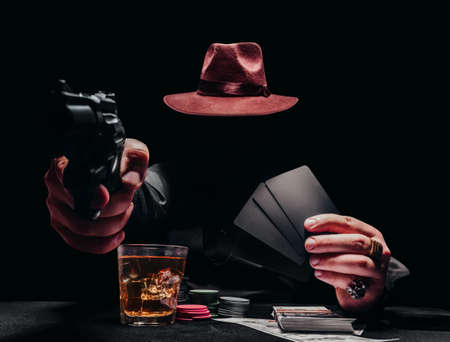 Photo of a shaded gangster pointing gun in black shirt and hat playing poker and holding black...
