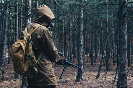 Photo of a nuclear war stalker soldier standing and posing in soviet gas mask, backpack with rifle in woods back view.の写真素材