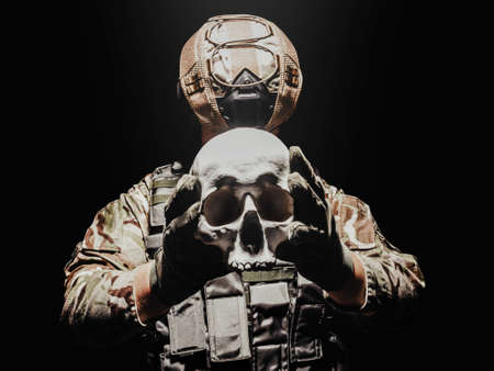 Photo of a fully equipped soldier in armor vest, helmet and gloves holding human skull.の写真素材