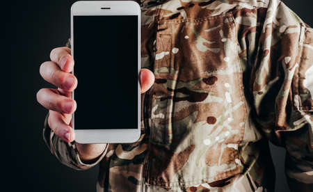 Photo of soldier in camouflaged uniform holding smartphone on black background.