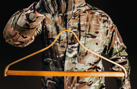 Photo of soldier in camouflaged uniform and tactical gloves holding wooden shirt hanger on black...