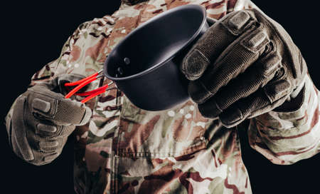 Photo of soldier in camouflaged uniform and tactical gloves holding metal hiking cup on black background.の写真素材