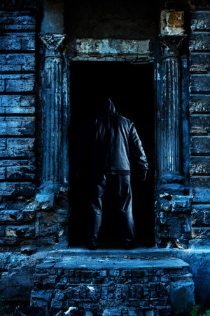 Photo of blue toned abandoned old and cracked building door entrance with hooded stalker man.の写真素材