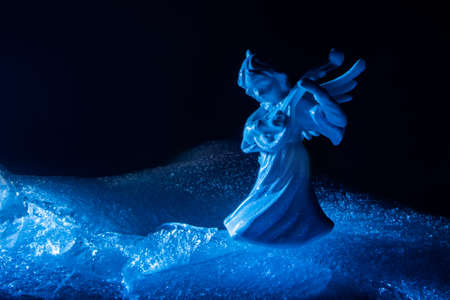 Photo of blue toned ice angel miniature playing the lute and standing on cracked ice surface on dark backdrop.の写真素材