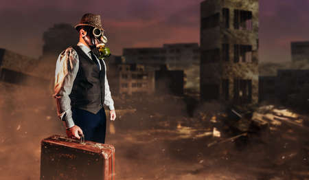 Photo of post apocalyptic male citizen in formal outfit, hat, gas mask and suitcase standing on destroyed city nuclear war wasteland.の写真素材
