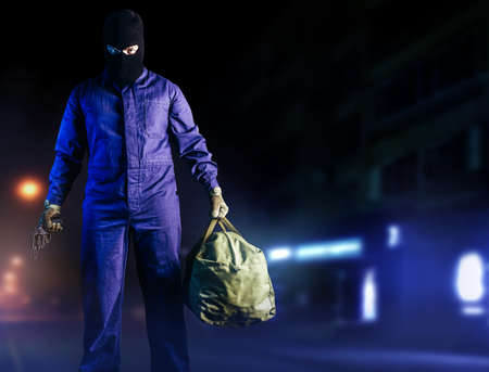 Photo of robber in mask, overalls, gloves, bag and gun standing front view on night street background.の写真素材
