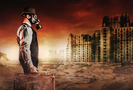 Photo of post apocalyptic male citizen in formal outfit, hat, gas mask and suitcase standing on destroyed city nuclear war wasteland.の写真素材