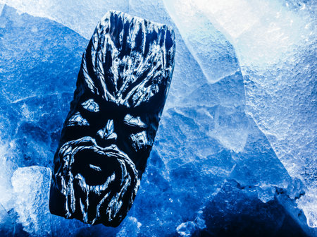 Photo of blue toned ancient wooden fantasy face idol totem laying on cracked ice surface.の写真素材