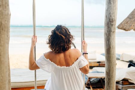 girl in a white dress swinging on a swing his back on the beach.の写真素材