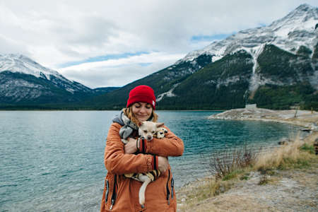 girl in an orange jacket holds a chihuahua in her arms near lake, canada.