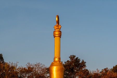 A tall gold statue with a golden top sits on a hill. The sky is blue and the sun is settingの写真素材