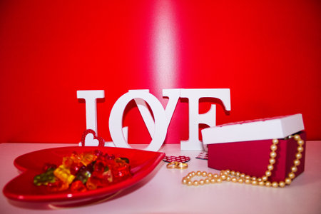 red heart-shaped plate with teddy bear colors jelly beans, word love in the background on a red background and pearl necklace in a gift boxの写真素材