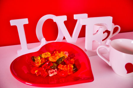 red plate in the shape of a heart with gummy bears on a red background and white letter forming the word love and two white cups with red heartの写真素材