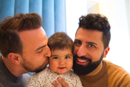 gay couple and their daughter sitting on the sofa watching television. The girl watches the cartoons attentivelyの写真素材